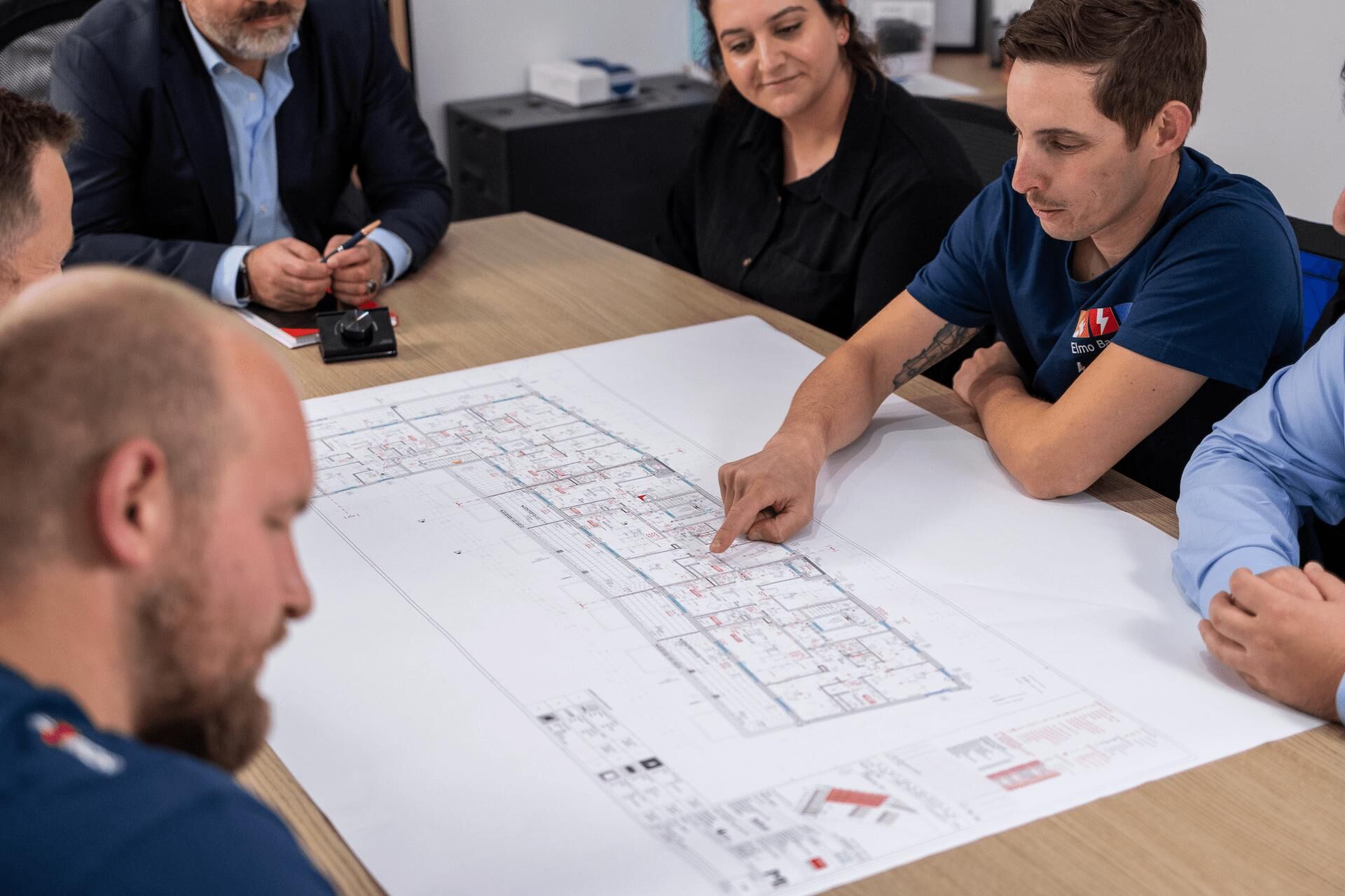 Gebäudetechnik - Elmo Bau GmbH Ein Team von Fachleuten diskutiert Baupläne an einem Konferenztisch; ein Mitarbeiter zeigt auf eine technische Zeichnung mit detailliertem Grundriss.