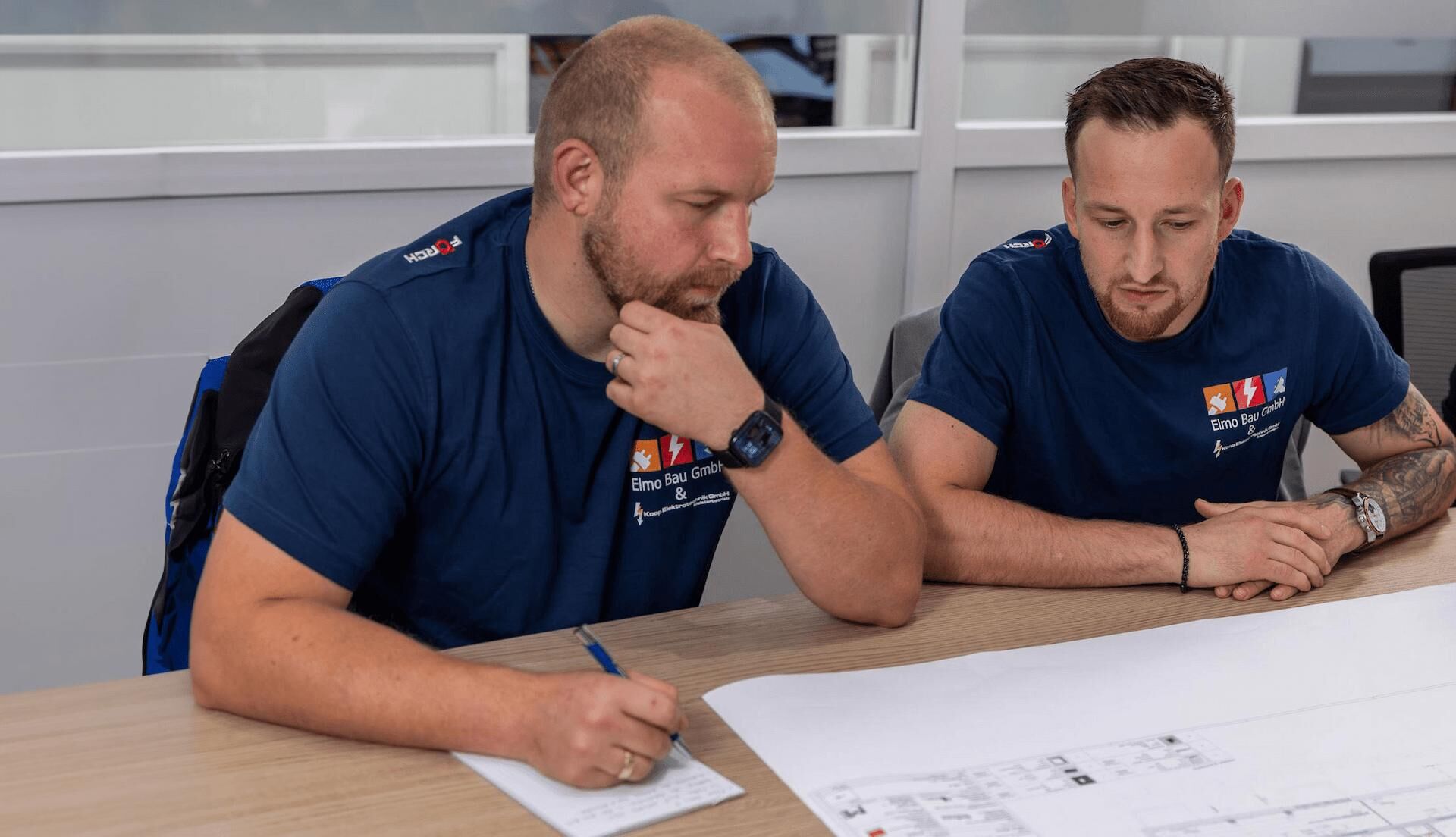 Your specialist for Electrical installation & cable laying Two men wearing blue Elmo Bau GmbH shirts are seated at a desk, attentively reviewing a large technical floor plan. One of them is taking notes with a pen, while the other is focused on the layout. Both appear concentrated and professional.