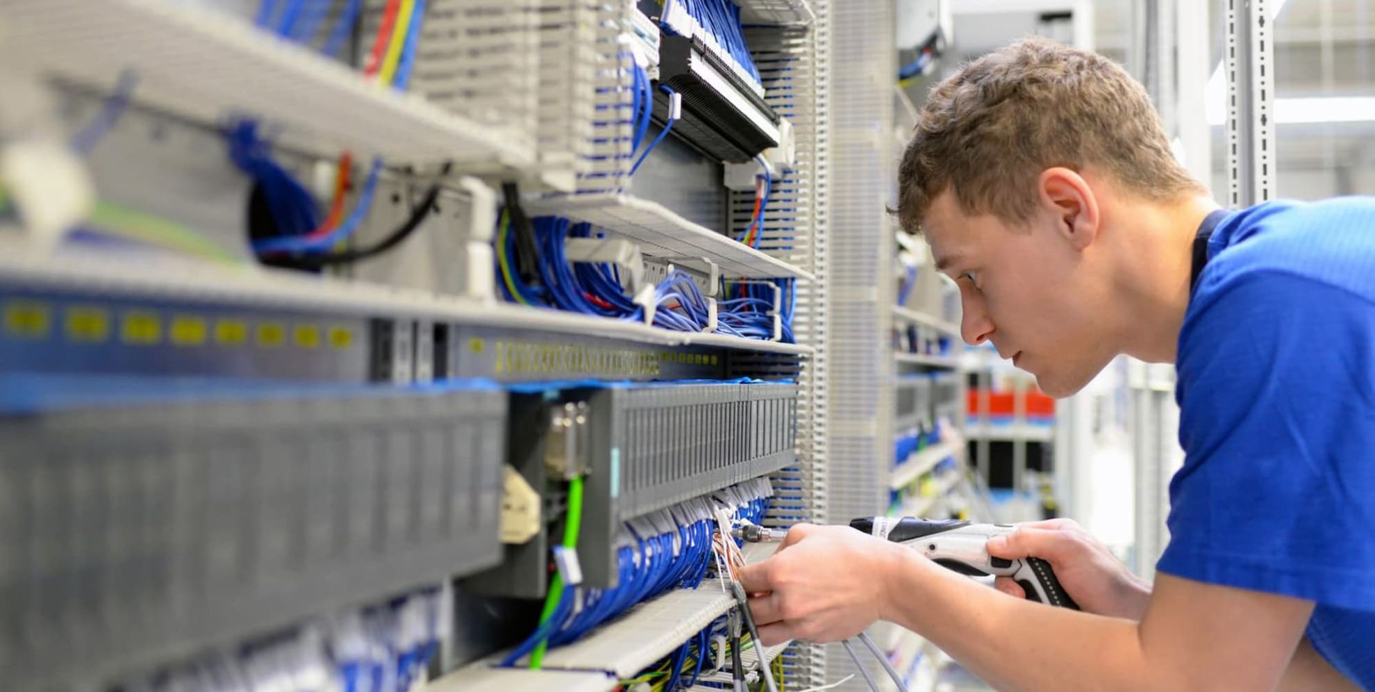 Bewirb dich jetzt für eine Ausbildung zum Elektroniker. Ein junger Mann im blauen T-Shirt arbeitet an einer Elektroanlage.