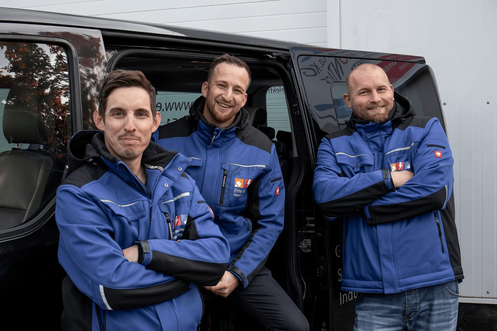 Elmo Bau Ihr Spezialist für Elektroinstallation & Kabelverlegung Drei Mitarbeiter der Elmo Bau GmbH in blauer Arbeitskleidung lächeln vor dem Firmenfahrzeug in die Kamera.