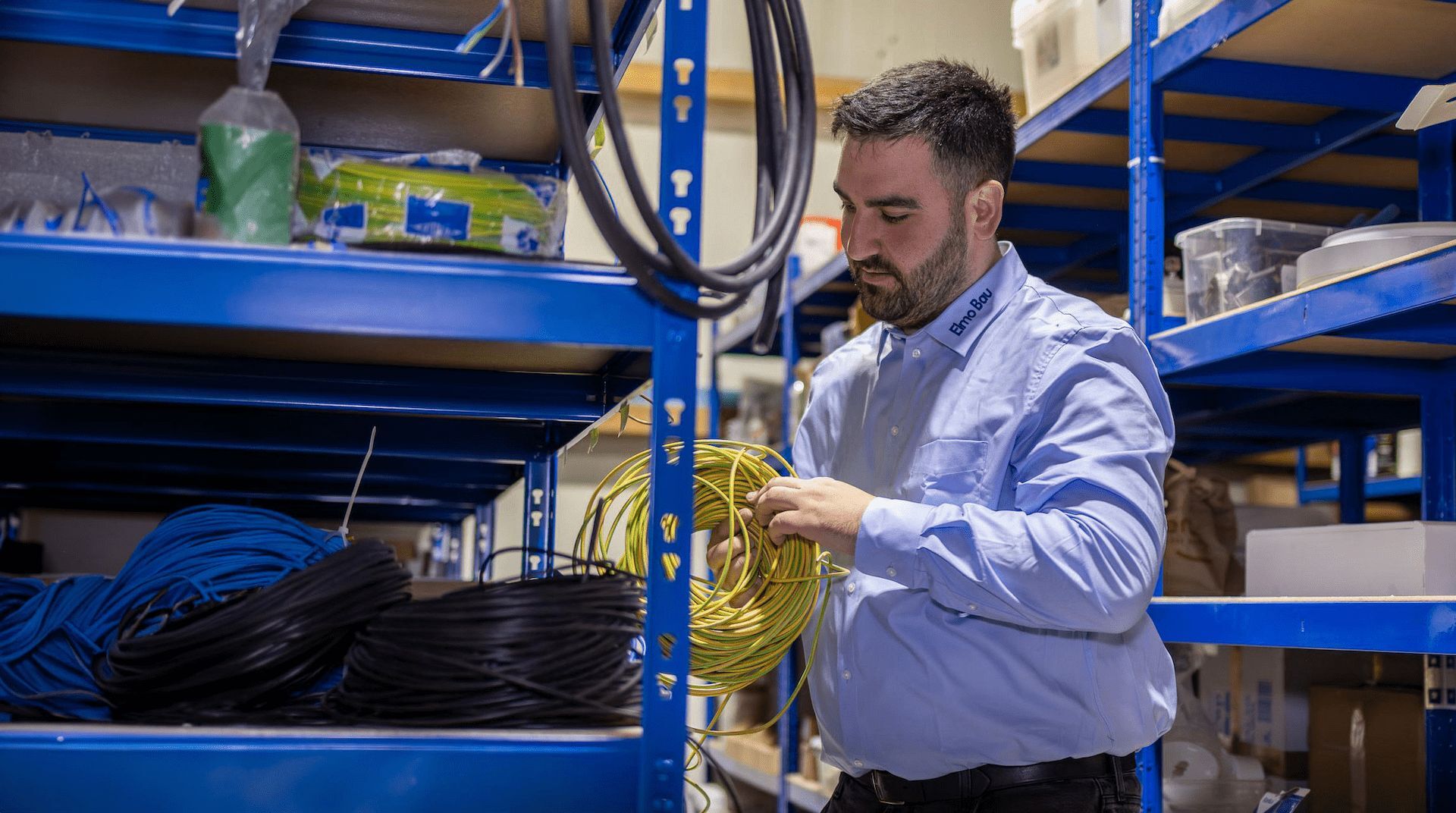 Elektroinstallation - Elmo Bau GmbH Ein Mitarbeiter in einem Lager arbeitet konzentriert mit einer Rolle gelben Kabels, umgeben von blauen Metallregalen mit verschiedenen Kabeln und Materialien.