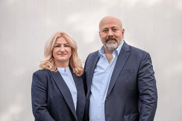 Porträt eines Mannes und einer Frau im Business-Outfit vor hellem Hintergrund. Beide tragen blaue Anzüge, die Frau hat blonde Haare, der Mann eine Glatze mit grauem Bart.