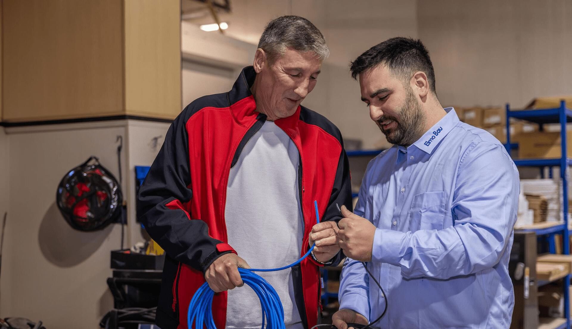 Zwei Männer stehen in einem Lagerraum und begutachten gemeinsam Kabel. Einer trägt ein Hemd mit dem Schriftzug „Elmo Bau“, der andere eine rote und schwarze Arbeitsjacke. Im Hintergrund sind Regale mit Arbeitsmaterialien und Werkzeugen zu sehen.