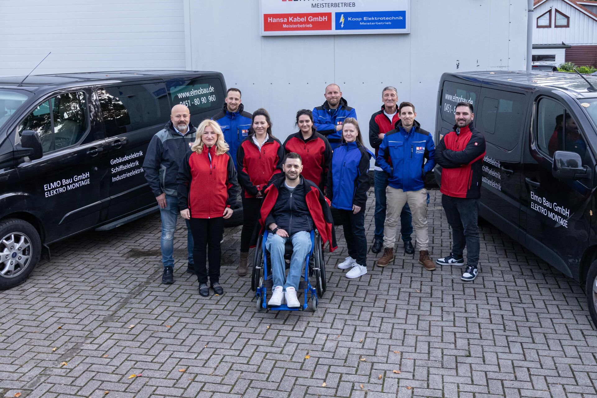 Unser Team - Elmo Bau GmbH Group photo of the Elmo Bau GmbH team with employees in red and blue work clothes, standing between two company vehicles, taken in front of the company building.