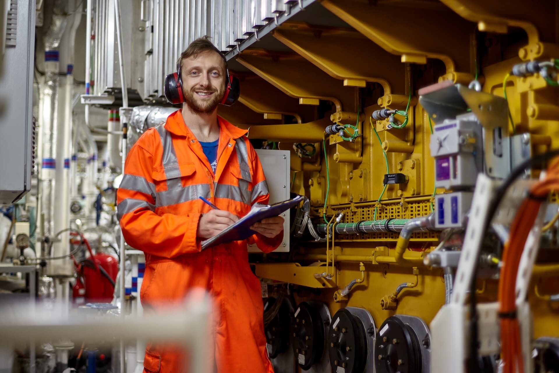Bewirb dich jetzt als Schiffselektriker! Porträt eines jungen Ingenieurs mit einem Lächeln in einem orangefarbenen Overall, der im Maschinenraum eine Checkliste abarbeitet.