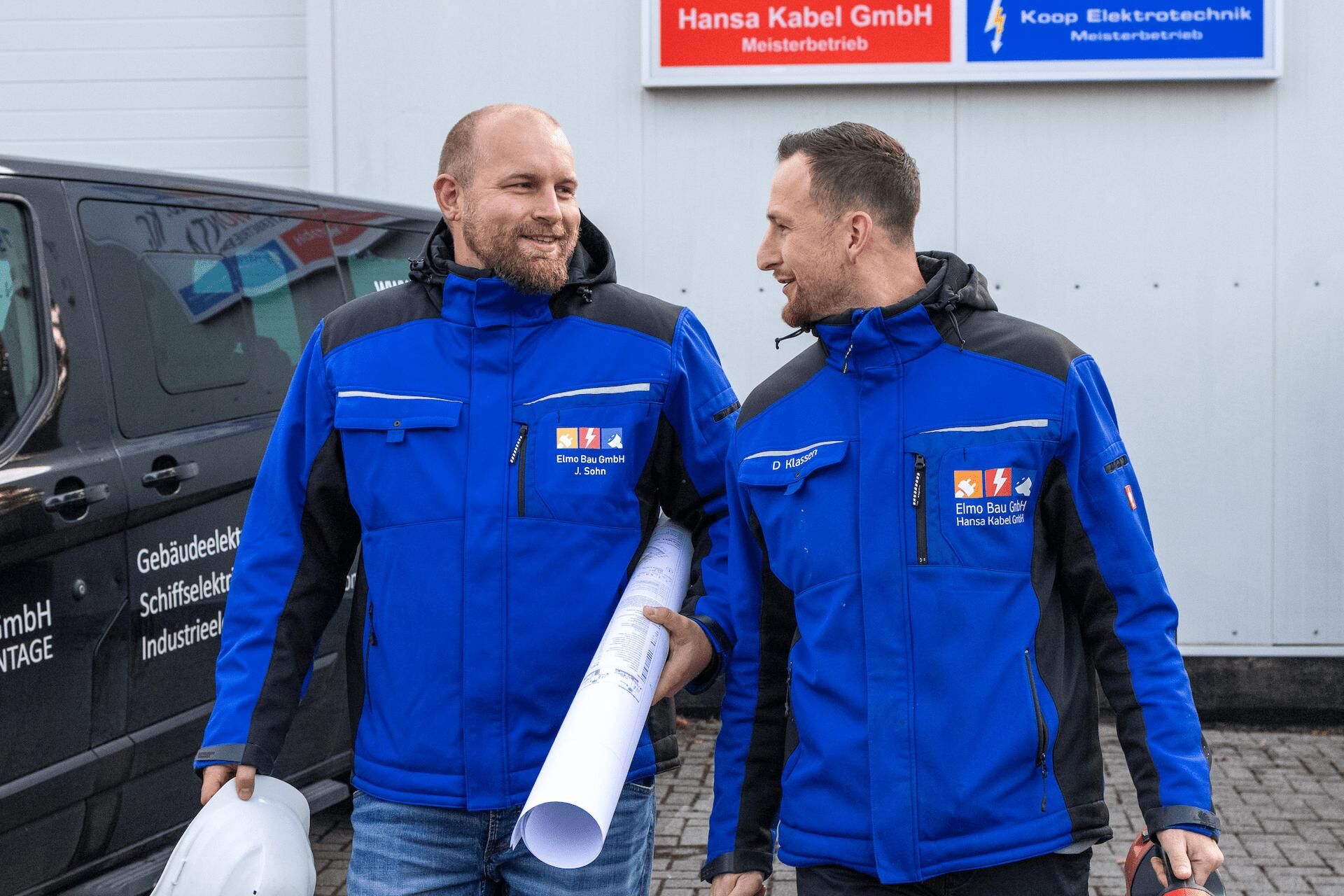 Elmo Bau Ihr Spezialist für Elektroinstallation & Kabelverlegung Zwei Elektriker in blauen Arbeitsjacken der Elmo Bau GmbH gehen lächelnd nebeneinander, einer trägt einen Bauhelm, der andere Baupläne. Im Hintergrund ist ein Firmenfahrzeug sowie ein Schild mit Logos zu sehen.